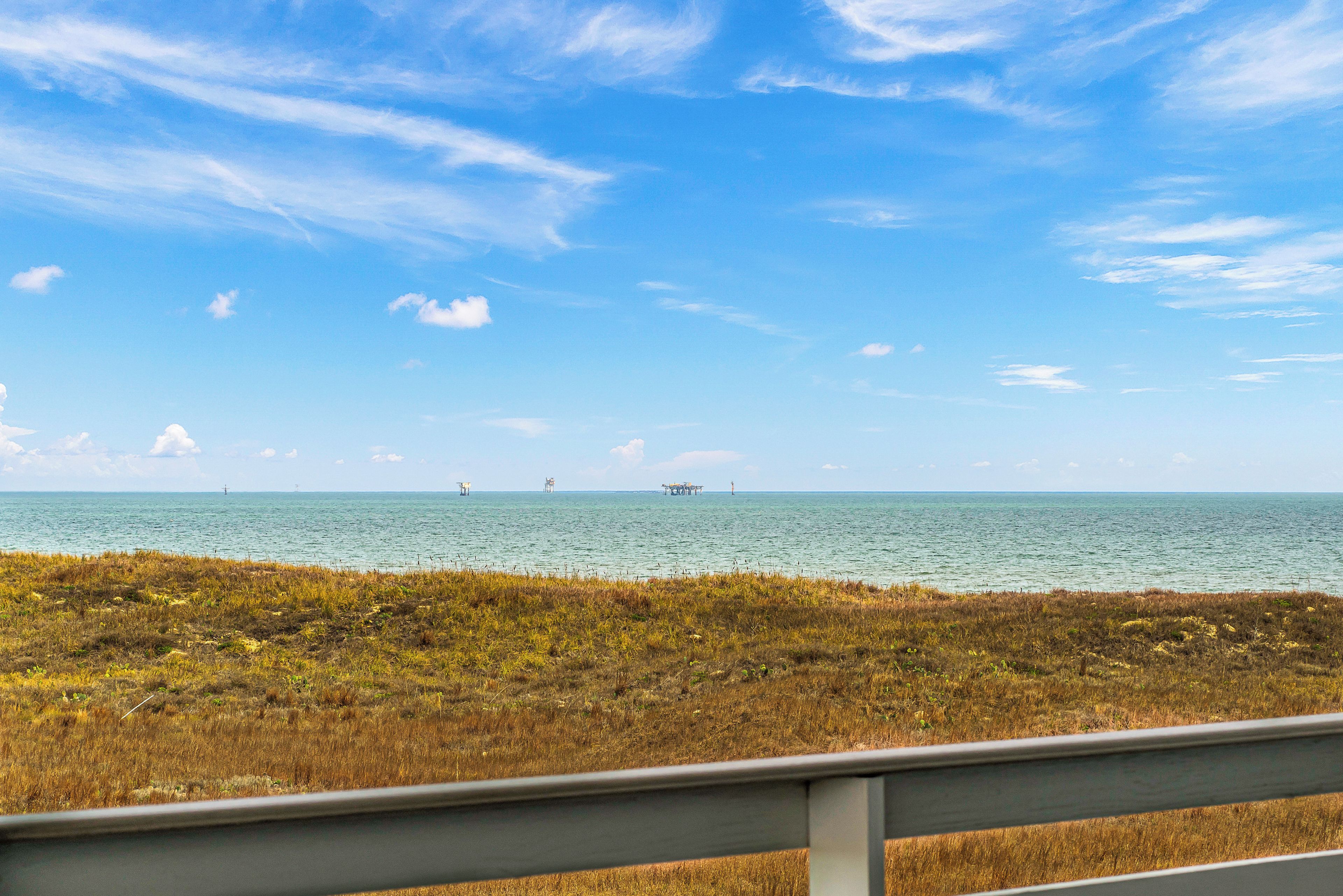 A coastal view with a grassy foreground, a clear blue sky, and distant offshore structures.