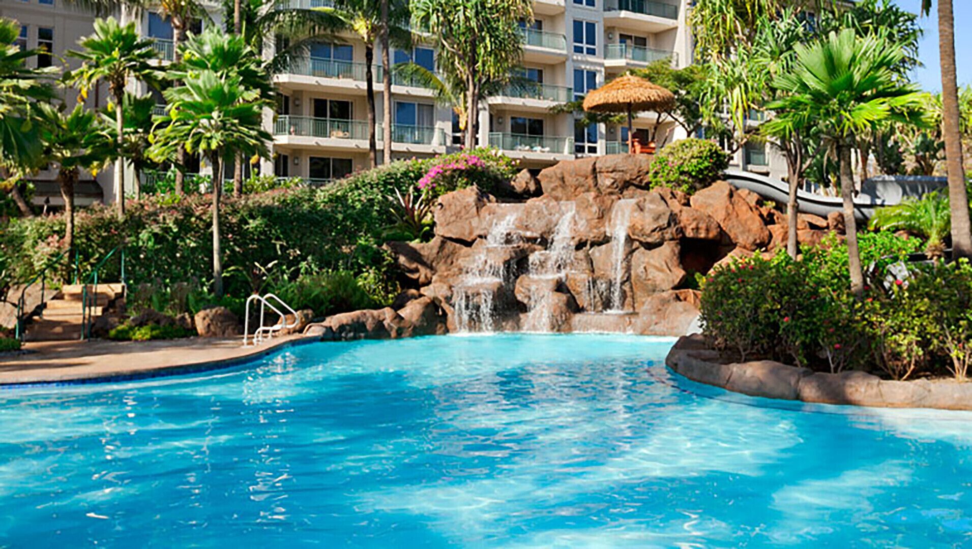 Piscina de hotel con cascada, rodeada de palmeras y un edificio de varios pisos con balcones.