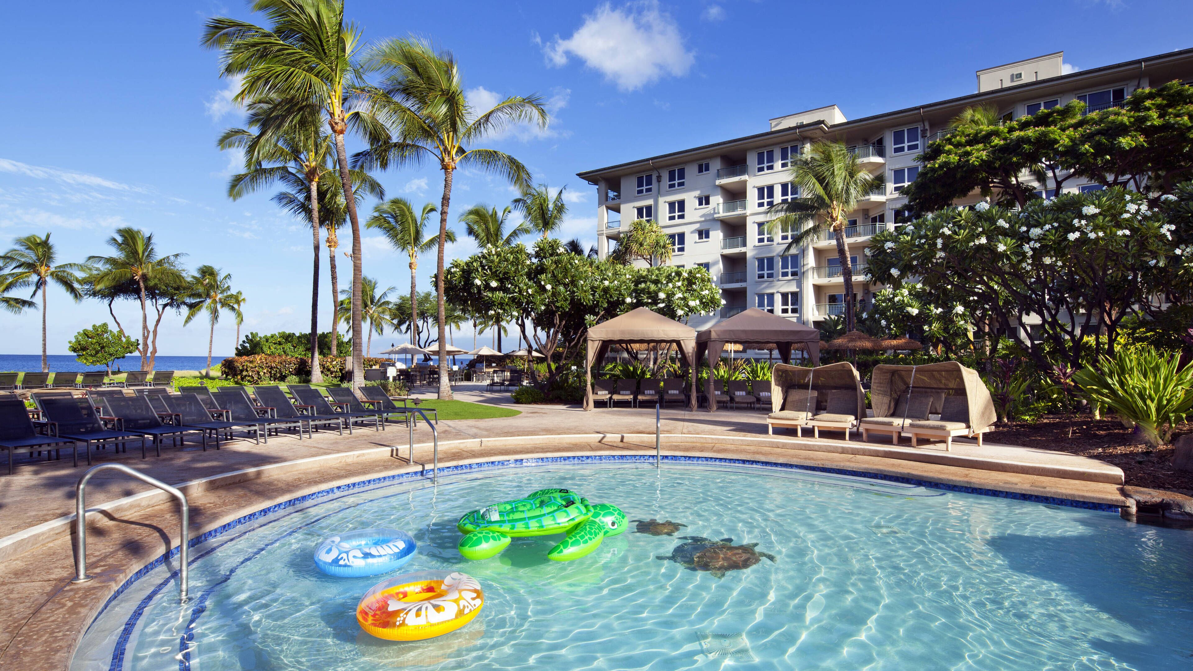 A pool area with lounge chairs, a small pool with inflatable toys, palm trees, and a building in the background.