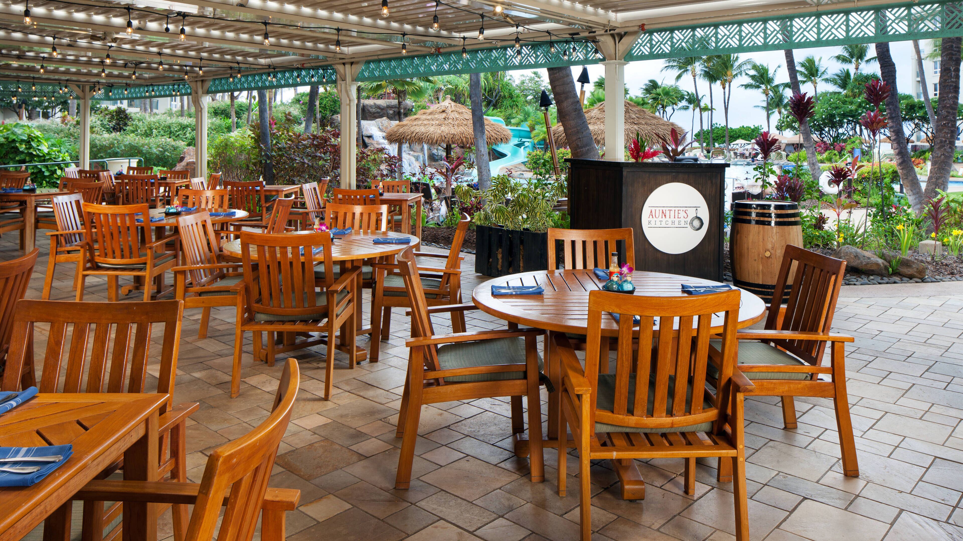 An outdoor dining area with wooden tables and chairs, a thatched-roof structure, and a view of a water feature and greenery.