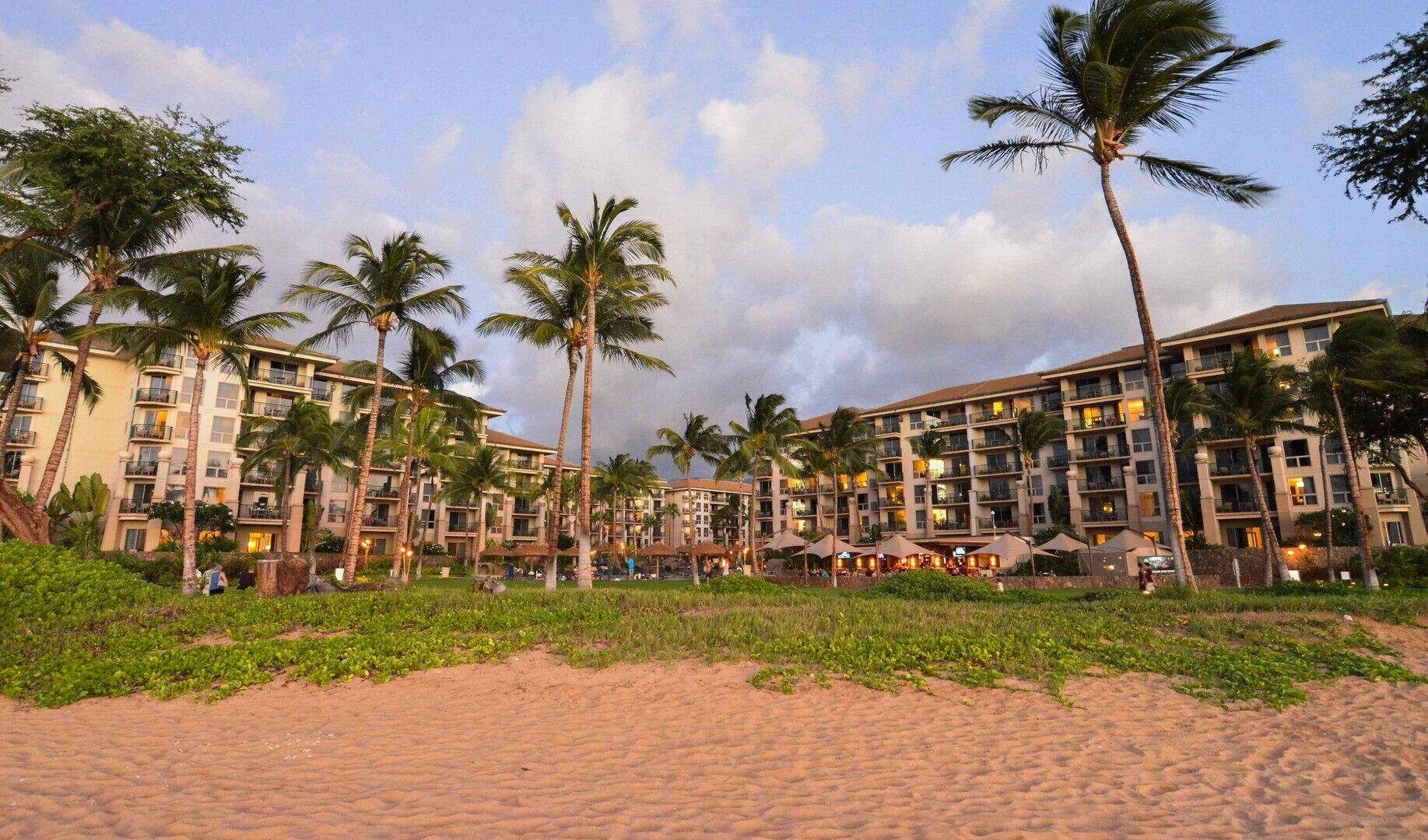Un hotel frente a la playa con varios pisos, palmeras y una zona de arena.