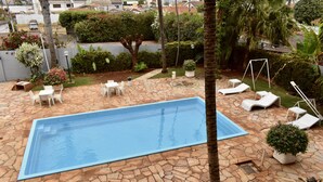A rectangular swimming pool with stone tiles, surrounded by white lounge chairs and tables, and various potted plants.