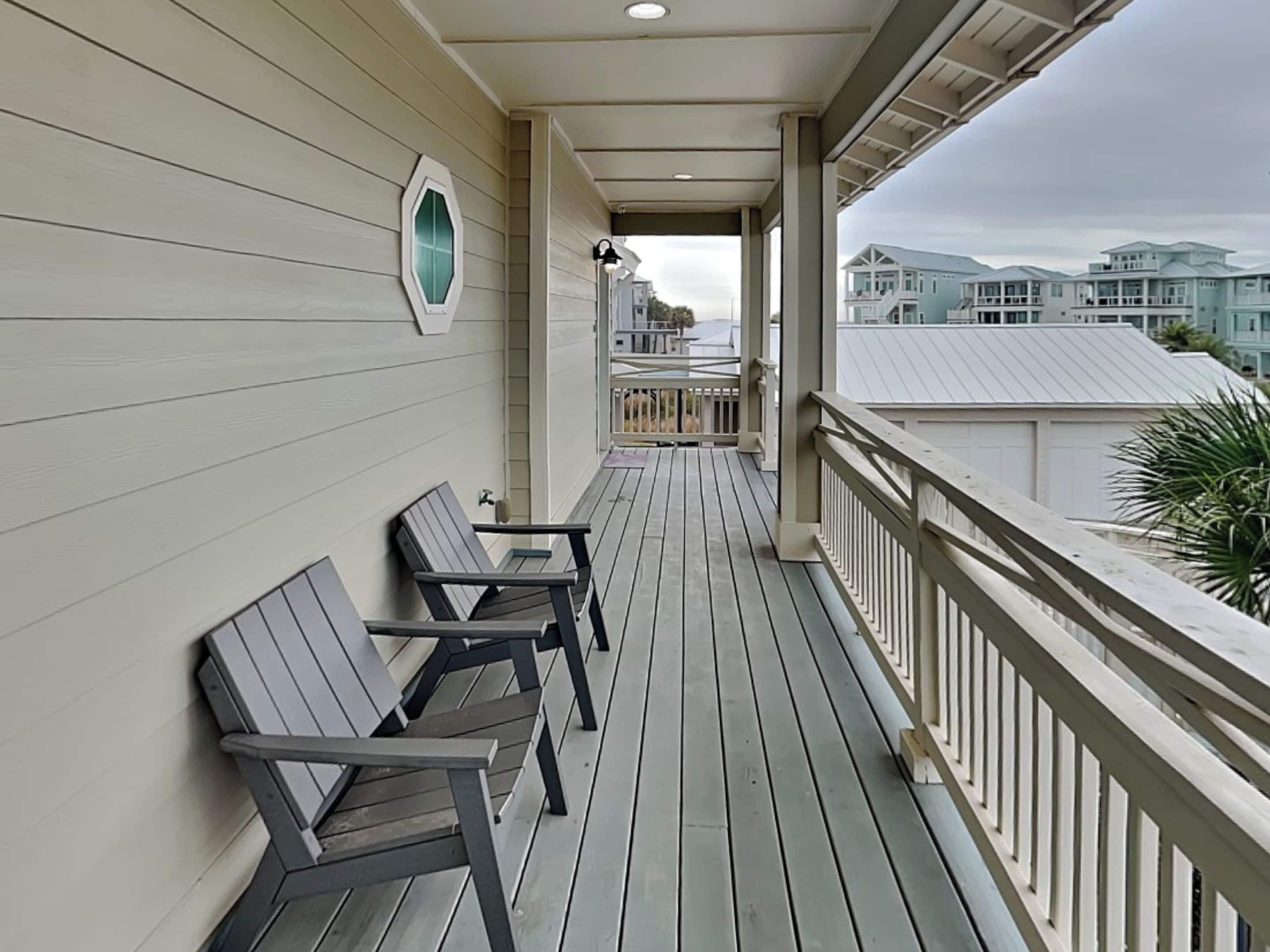 Une terrasse en bois avec deux chaises et une petite table, une balustrade blanche et une vue sur des maisons au loin.
