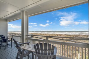 A covered deck with two Adirondack chairs and a table, overlooking a beach with a clear sky. - It'll Do by Silver Sands Vacation Rentals (Port Aransas)