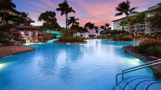 A resort pool with a waterfall, palm trees, and a building in the background.