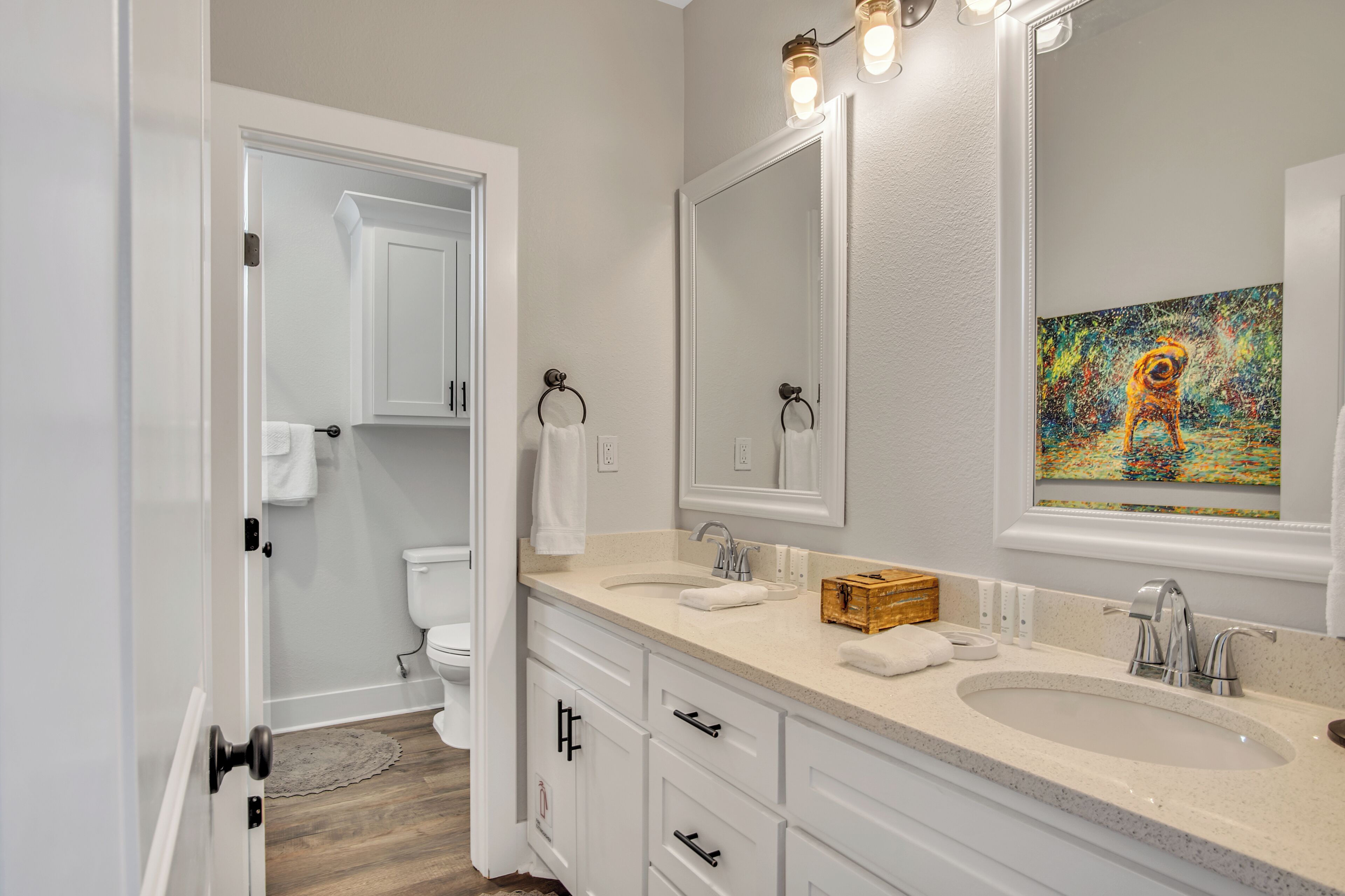 A modern bathroom with two sinks, a large mirror, and a painting above the mirror.