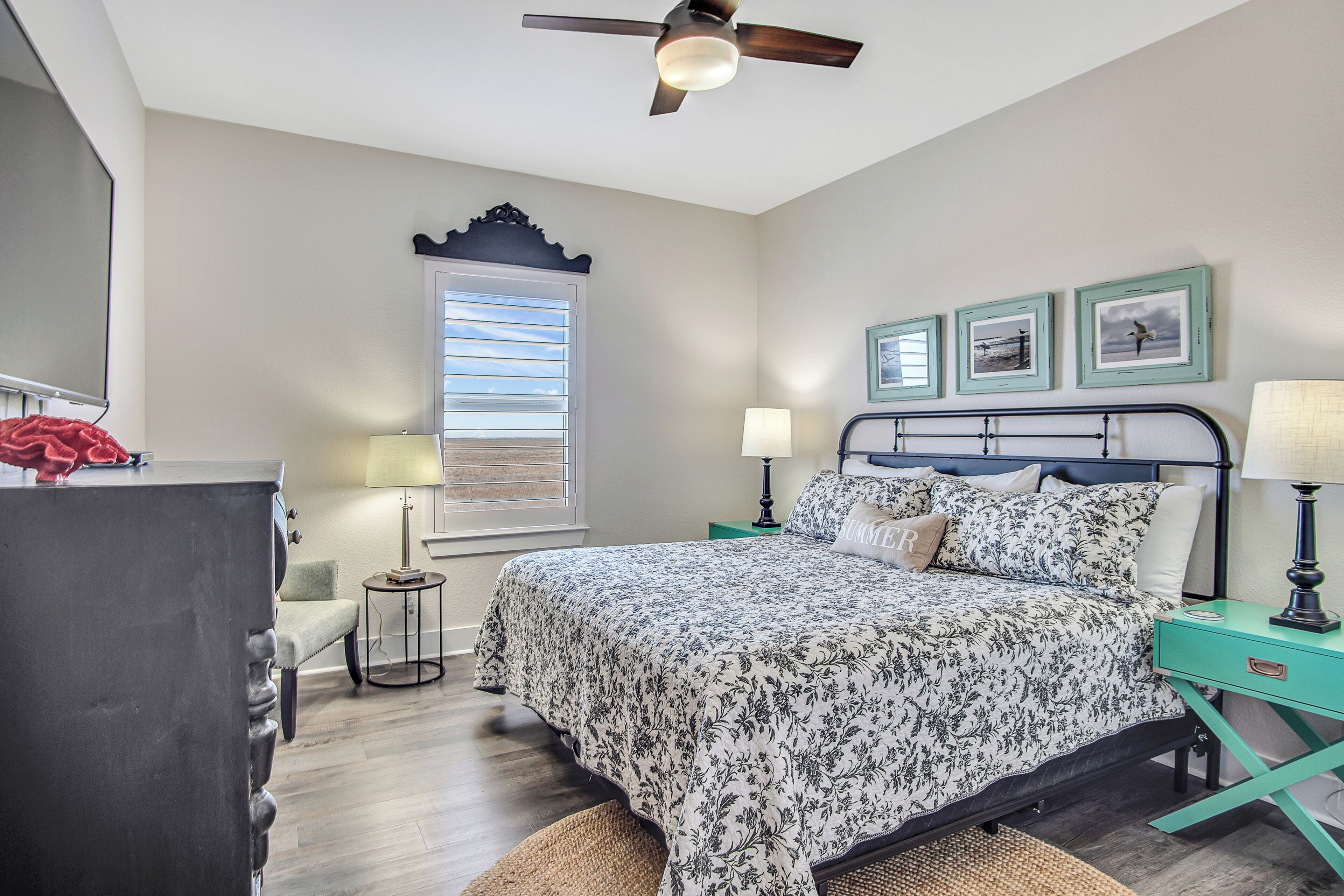 A bedroom with a bed, bedside tables, a ceiling fan, a window with blinds, and framed pictures on the wall.