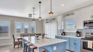 A modern kitchen with a central island, bar stools, and a view of the outside through large windows. - Downtime by Silver Sands Vacation Rentals (Port Aransas)