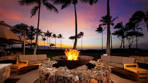 A stone fire pit with a fire in the center, surrounded by wicker chairs, set against a sunset with palm trees in the background.