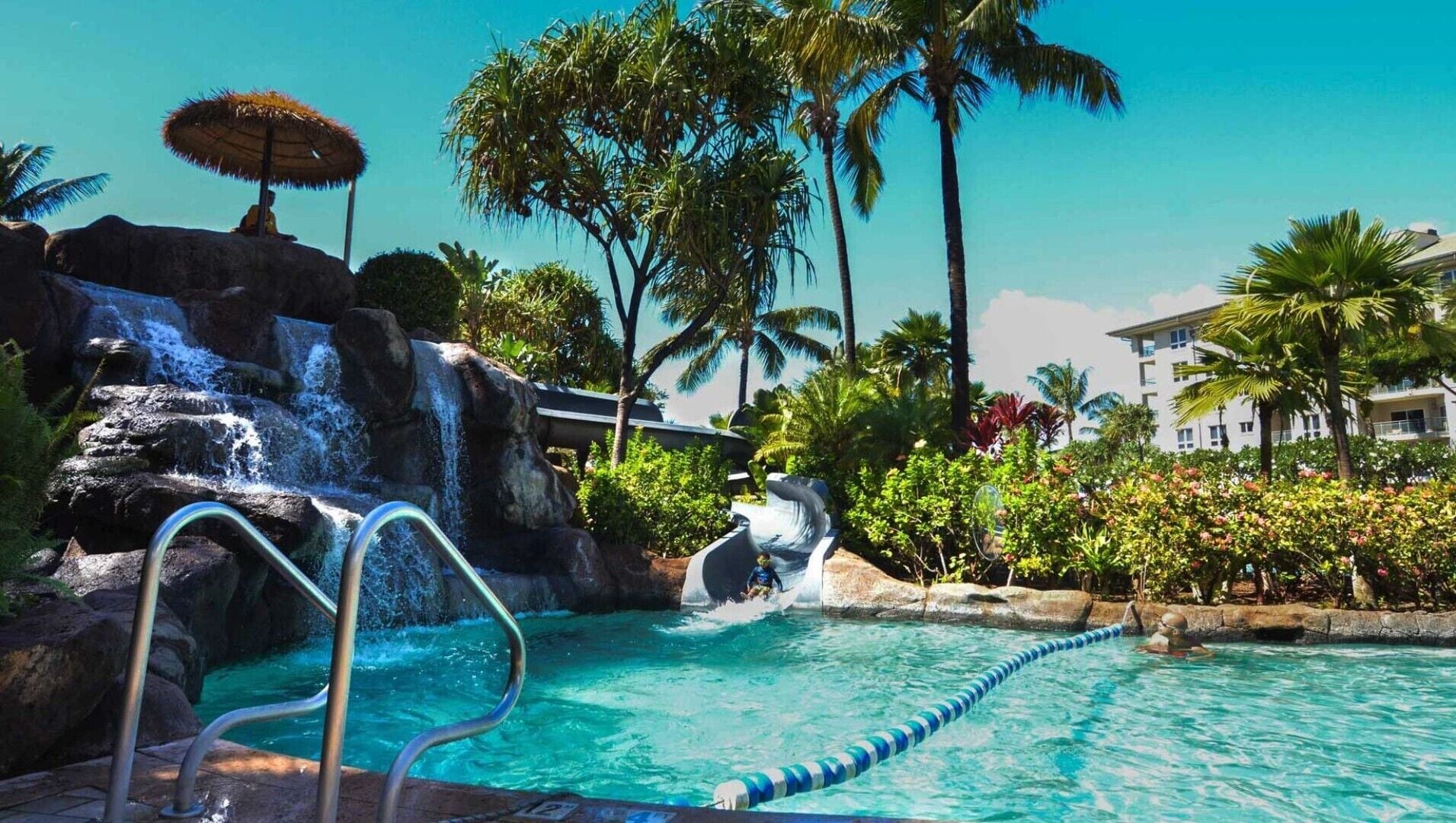 En swimmingpool med et vandfald, omgivet af palmer og en bygning i baggrunden.