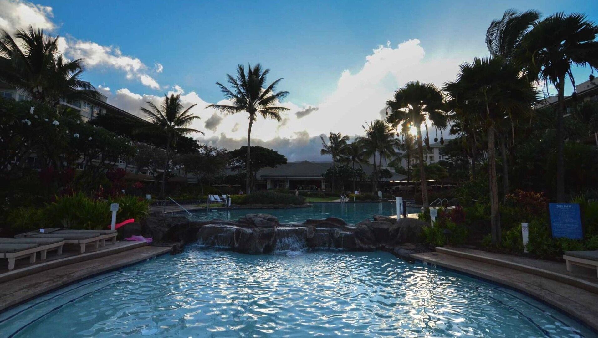 En pool på ett semesterhotell med en konstgjord vattenfallskaskad, omgiven av palmer och solstolar.