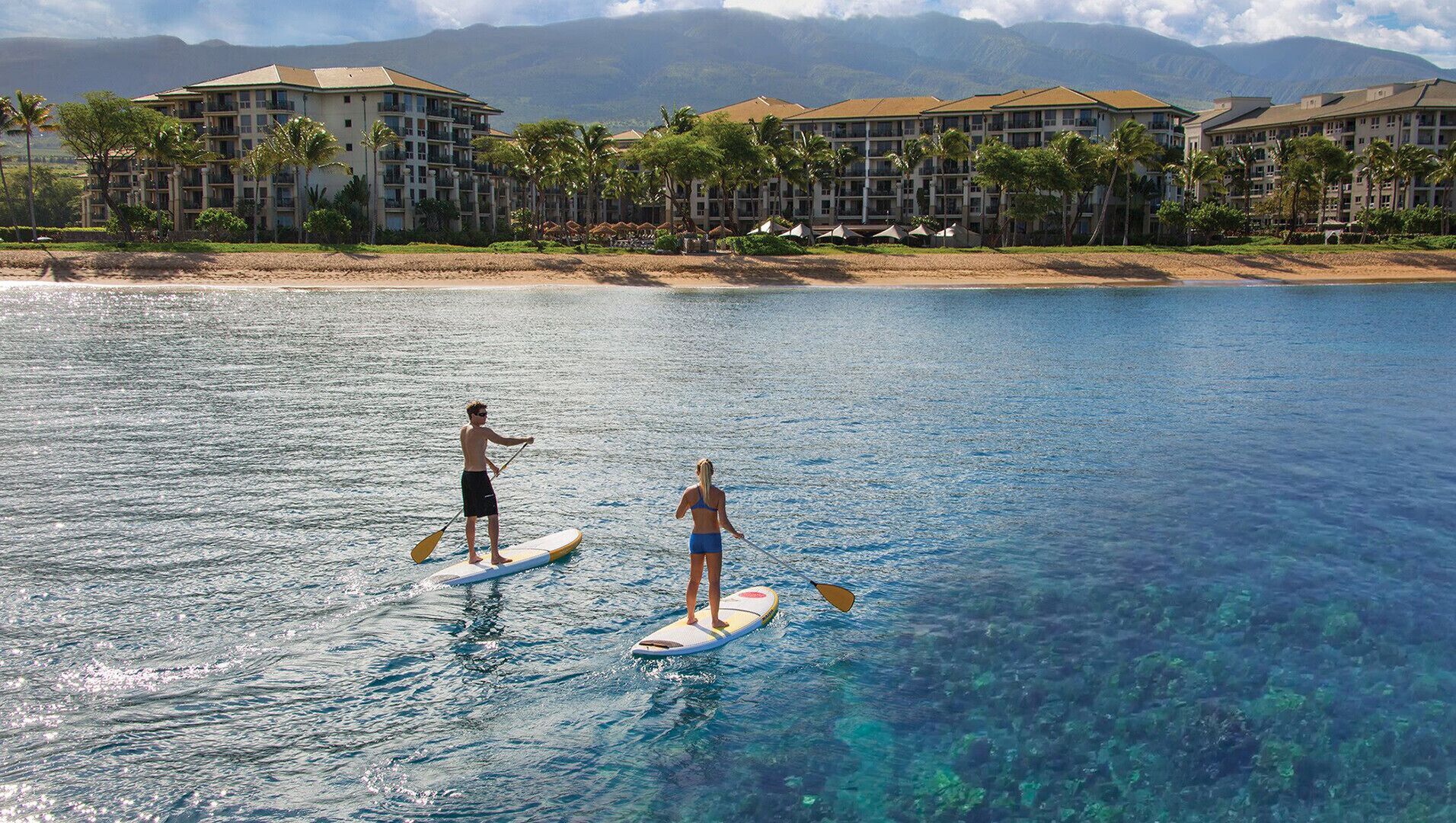 Zwei Personen stehen auf Paddleboards auf einem klaren, blauen Meer, im Hintergrund ein Resort.