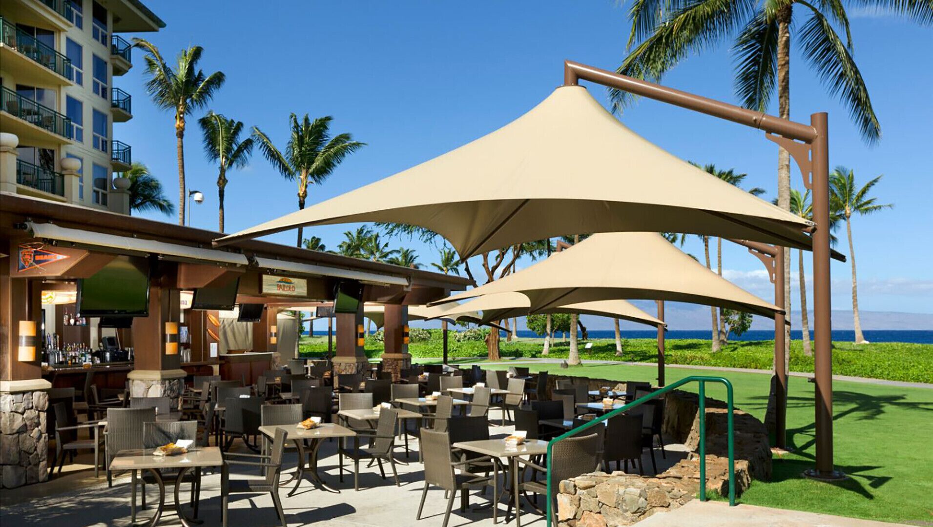 Un área de comedor al aire libre frente a la playa, con mesas y sillas, protegida por grandes sombrillas, y una zona de bar con vistas al océano.