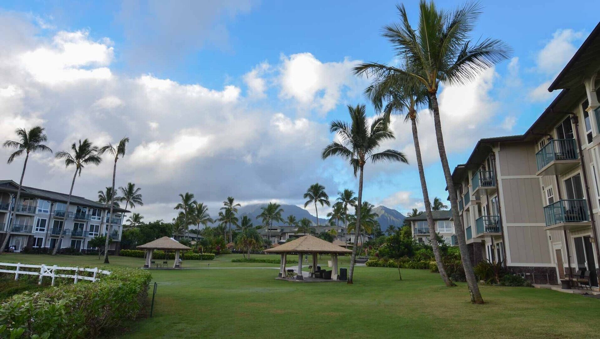 Un resort con edificios de varios pisos, palmeras y un área verde con dos pabellones.