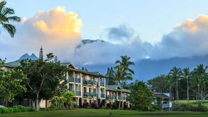 A multi-story hotel with balconies, surrounded by lush greenery and palm trees, with a mountain and clouds in the background.