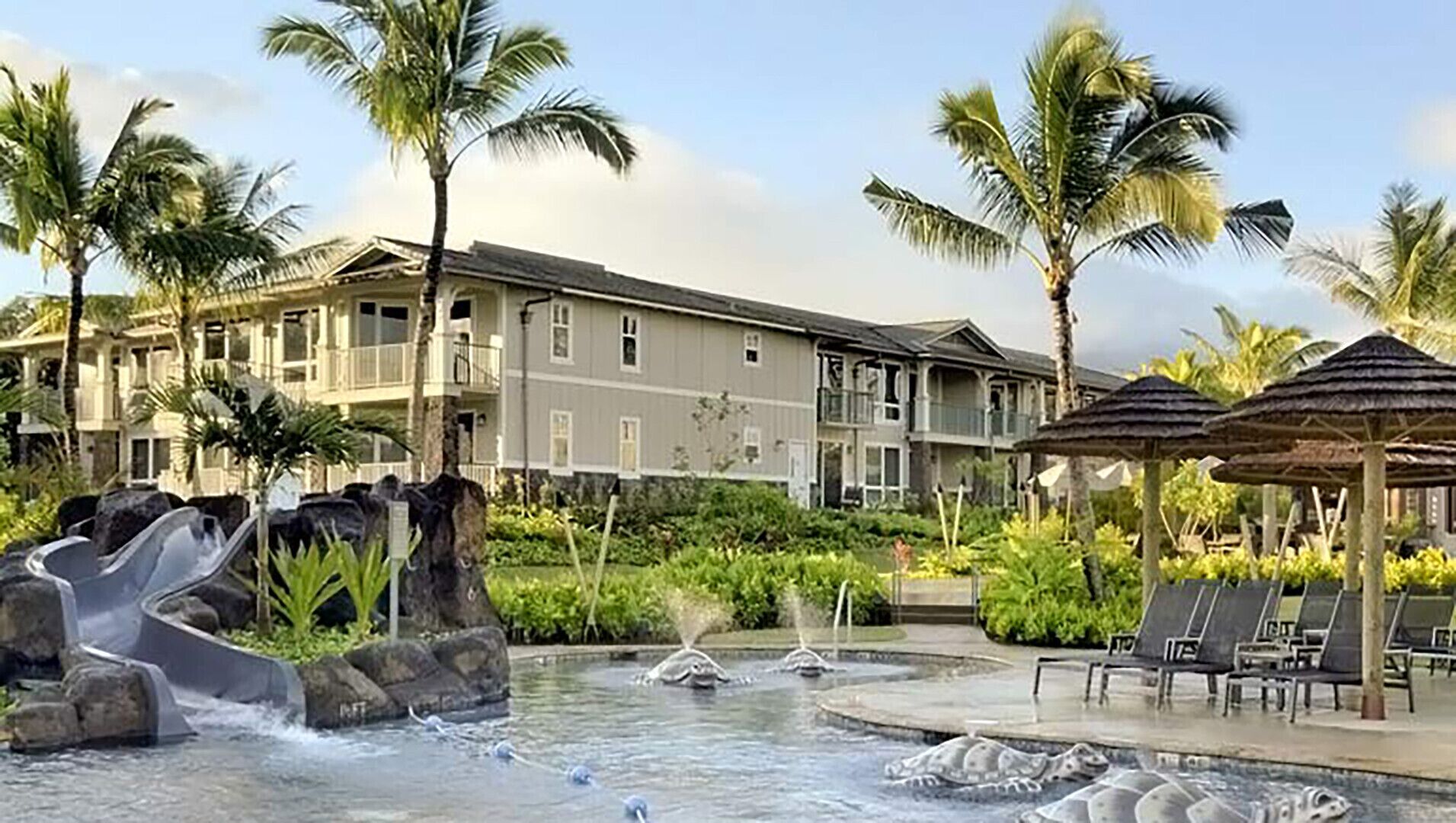 A resort with a pool area featuring water slides and a fountain, surrounded by palm trees and lounge chairs.