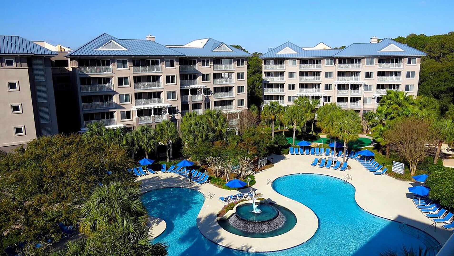 Un hotel con un amplio espacio de piscina, que incluye una fuente central, rodeado de sillones y sombrillas.