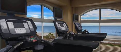 A gym with treadmills and exercise equipment, large windows with a view of the ocean.