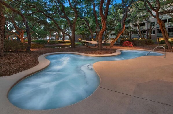 Una piscina con diseño ondulado, rodeada de árboles y un edificio al fondo.