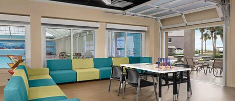 Um espaço de escritório moderno com assentos em tons de verde-água e amarelo, uma mesa branca com um jogo de tabuleiro e grandes janelas com vista para o exterior.