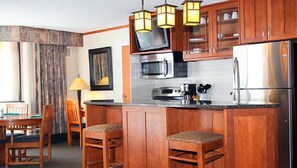 Cocina compacta con armarios de madera, microondas, estufa y refrigerador. Hay dos taburetes de bar y una mesa de comedor con sillas.