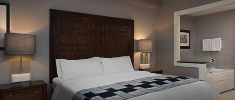 A hotel room with a large bed, two bedside tables with lamps, a dresser, a bathroom with a sink and mirror, and a framed picture on the wall.