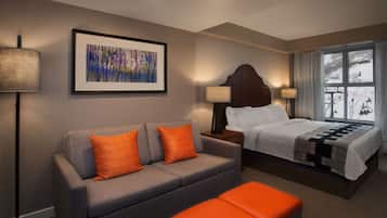 A hotel room with a grey sofa, two orange cushions, a bed with white bedding, a large window with a view of a snowy landscape, and a framed abstract painting on the wall.