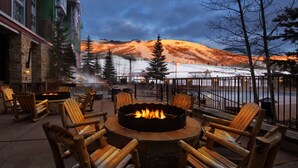 A fire pit with chairs around it, overlooking a snowy landscape with trees and a building in the background.