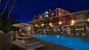 A swimming pool with lounge chairs and umbrellas, surrounded by a building with red exterior and string lights.