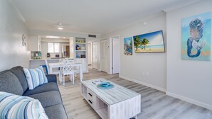 A modern living room with a gray sofa, a white coffee table, and a dining area with white chairs. There is a painting of a mermaid on the wall.