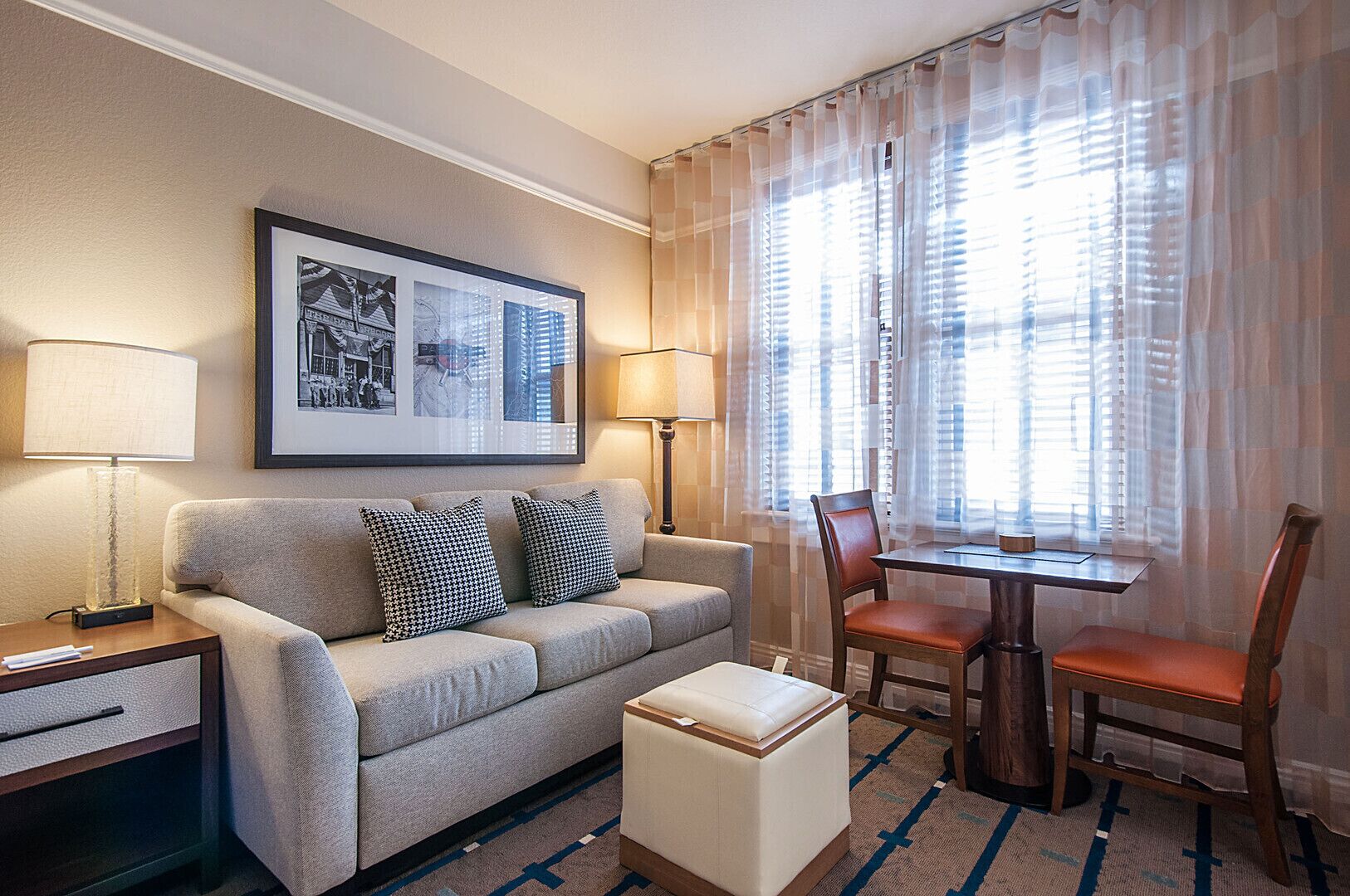 Habitación de hotel con sofá, dos almohadas, una mesa con sillas, una lámpara y un cuadro en la pared.