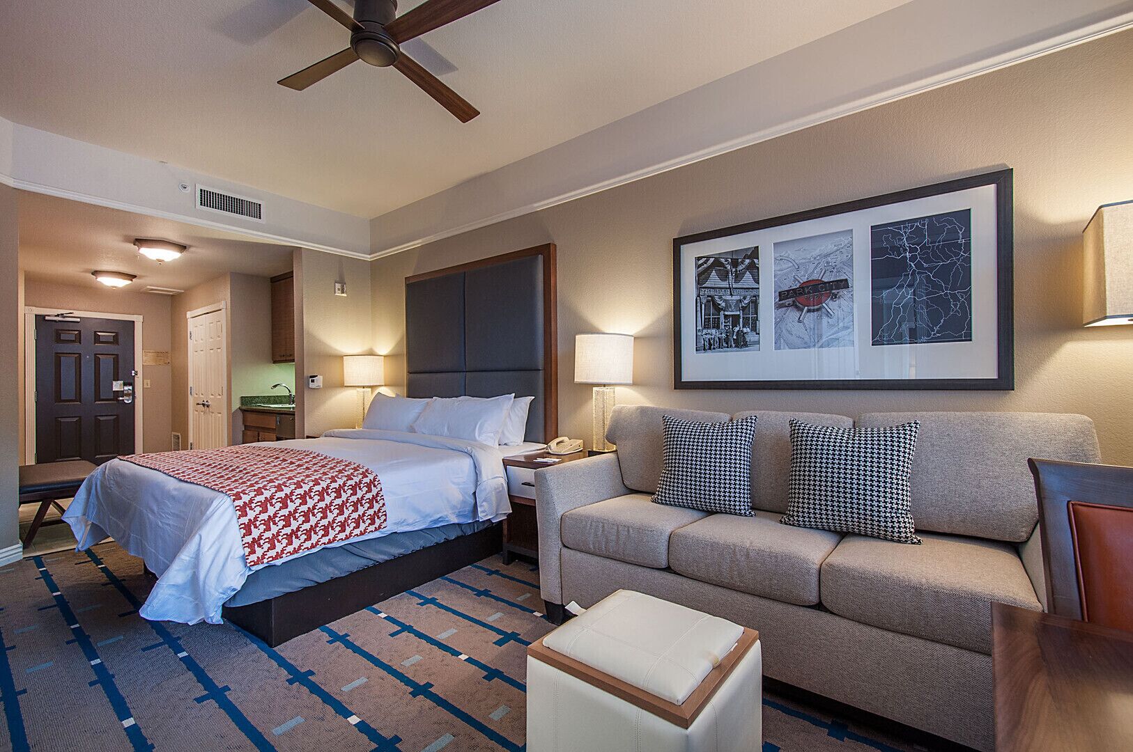 A hotel room with a bed, a sofa, a small table, and a ceiling fan. There is a framed picture on the wall and a door leading to another room.