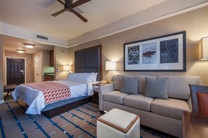 Une chambre d’hôtel avec un lit, un canapé, une petite table et un ventilateur de plafond. Un tableau encadré est accroché au mur, et une porte donne accès à une autre pièce.