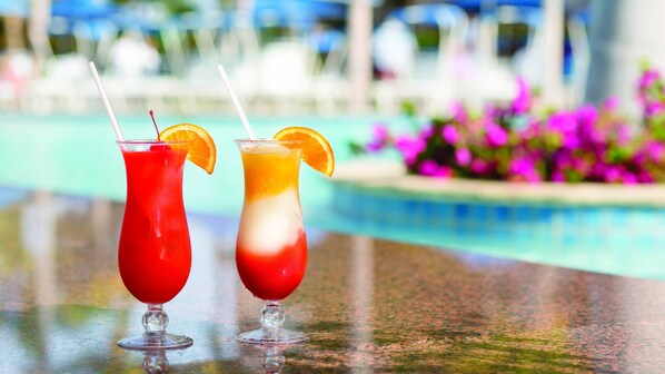 Two cocktails with straws and orange slices on a bar counter near a pool.