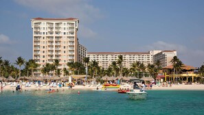 Un hôtel situé en bord de mer, avec plusieurs étages, une plage de sable et des palmiers.