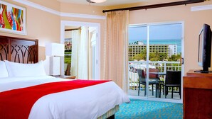 A hotel room with a bed, a TV, a balcony with a table and chairs, and a view of the ocean.