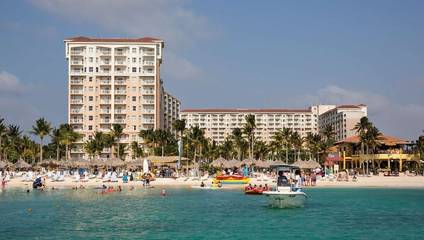 A beachfront hotel with multiple floors, palm trees, and a beach area with umbrellas and people.