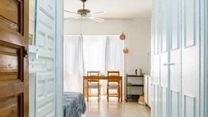 Una habitación con cama, una mesa de comedor con sillas, un ventilador de techo y una ventana con cortinas traslúcidas.