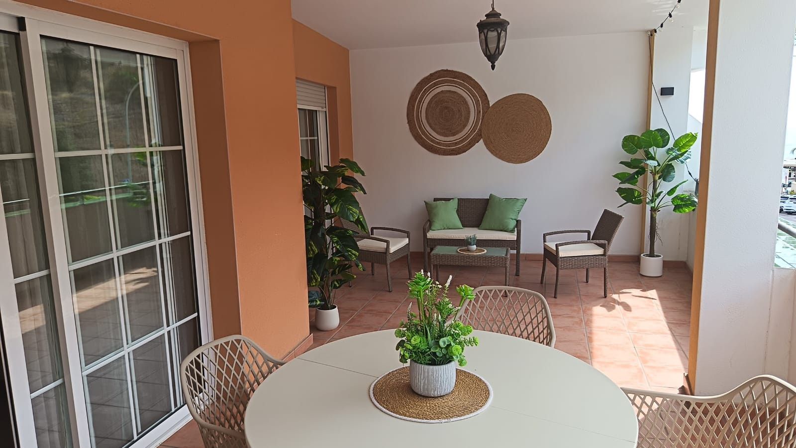 Un patio con una mesa redonda, sillas de mimbre, un sofá con cojines y plantas en macetas.