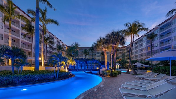 A resort pool area with a curved pool, palm trees, and lounge chairs.