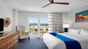 A hotel room with a bed, a desk, a chair, a TV, and a balcony with a view of the beach and ocean.