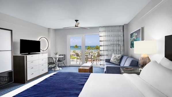 Une chambre d’hôtel comprenant un lit, une télévision, un bureau avec une chaise, un balcon avec vue sur la plage et un ventilateur de plafond.