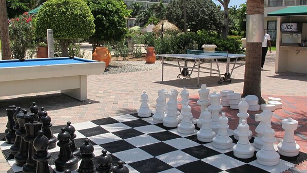 Un tablero de ajedrez con piezas dispuestas en dos filas, una mesa de billar y una mesa de tenis de mesa en un espacio al aire libre con plantas en macetas y un edificio al fondo.