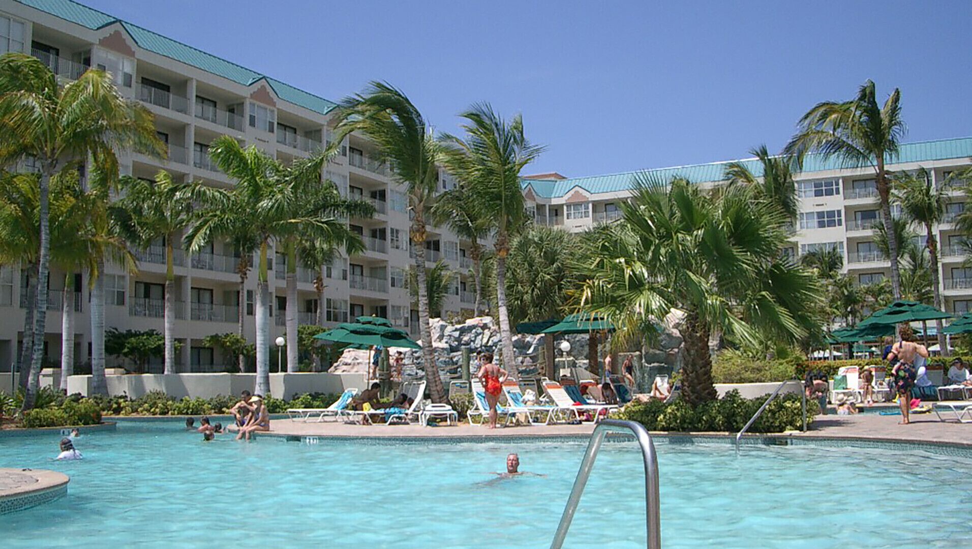 Un espace piscine d’hôtel doté de nombreuses chaises longues et de parasols, entouré de palmiers et d’un immeuble à plusieurs étages avec des balcons.