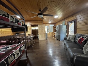 Interior de una cabaña con techo de madera, ventilador de techo, comedor, cocina con refrigerador y sala de estar con sofá y cojines estampados.