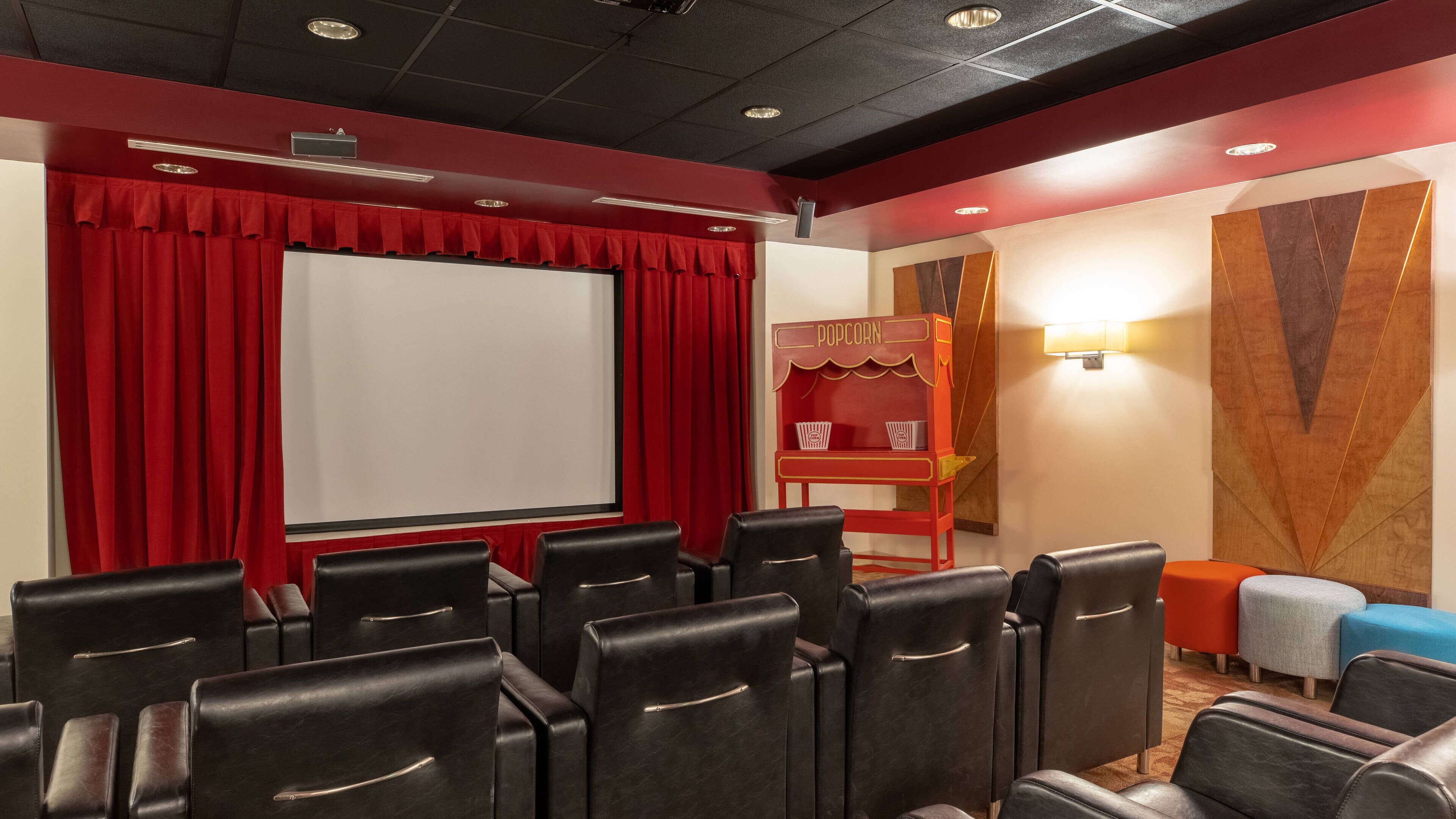Een theaterzaal met rode gordijnen, een wit scherm, zwarte leren stoelen, een popcornkraam en een muur met geometrische patronen.