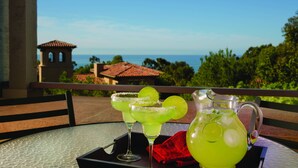 Een tafel met twee margarita’s en een karaf met een groene drank, op een balkon met uitzicht op de oceaan en een gebouw op de achtergrond.