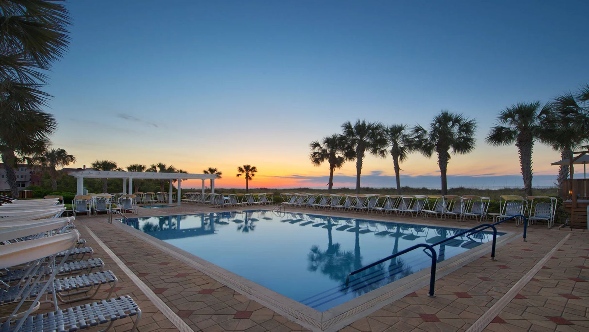 Une piscine bordée de chaises longues et de palmiers, le tout sur fond de coucher de soleil.