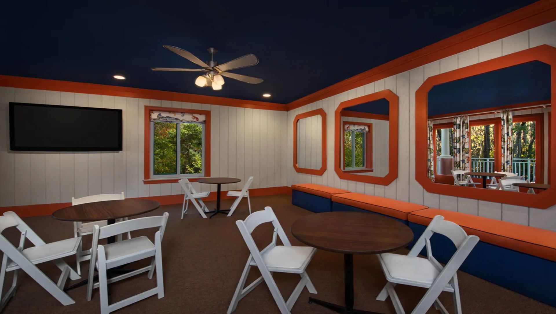 A room with white walls, orange trim, and a ceiling fan. There are several tables and chairs, and a large window with a view of trees.