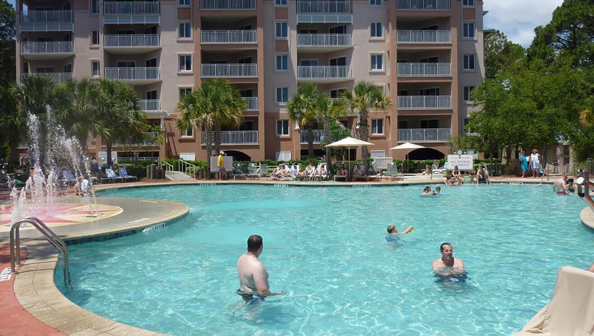 A swimming pool with people in the water, surrounded by a building with multiple balconies and palm trees.